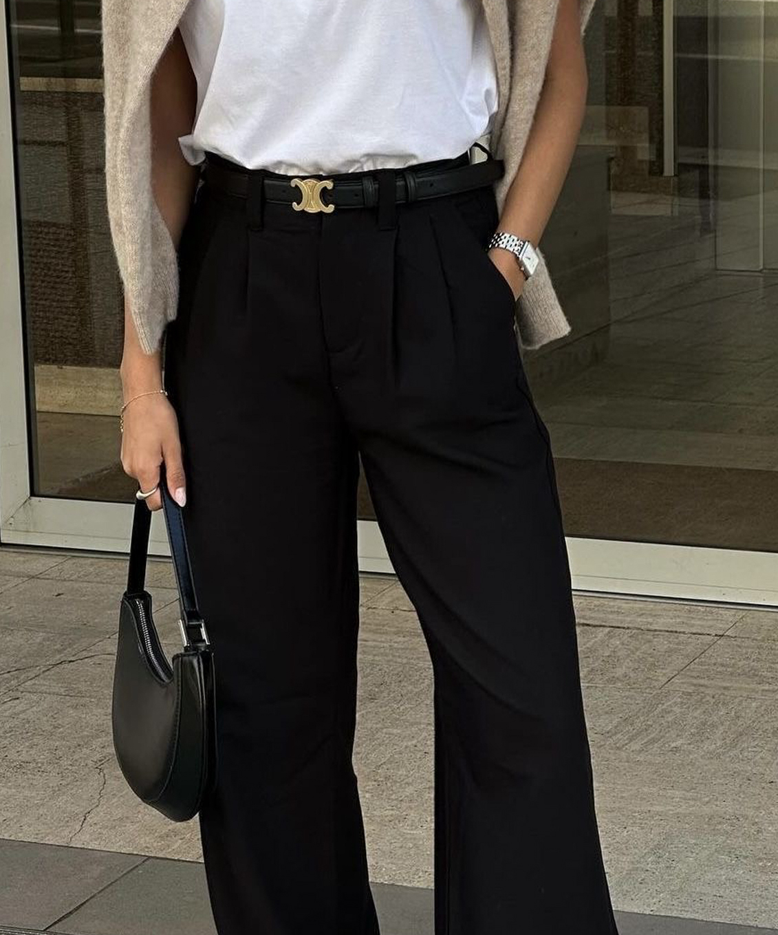 Femme portant un pantalon noir taille haute à jambes larges, un t-shirt blanc et une ceinture noire, avec un cardigan beige sur les épaules.