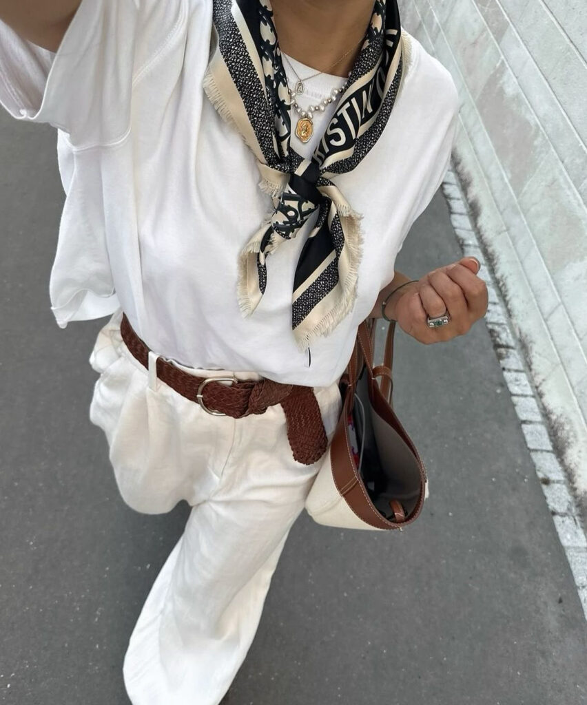 Femme portant un t-shirt blanc, un foulard noir et blanc noué au cou, un pantalon blanc et une ceinture brune tressée.