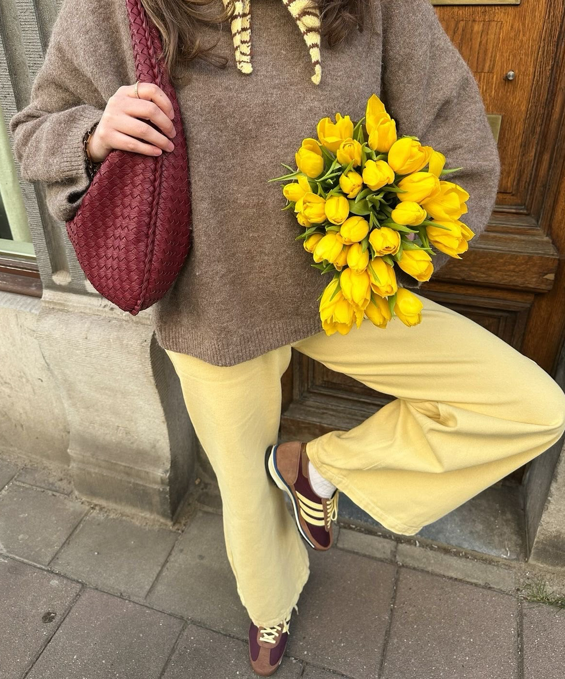 Femme portant un chandail brun et un pantalon jaune, tenant un bouquet de tulipes jaunes et un sac bourgogne