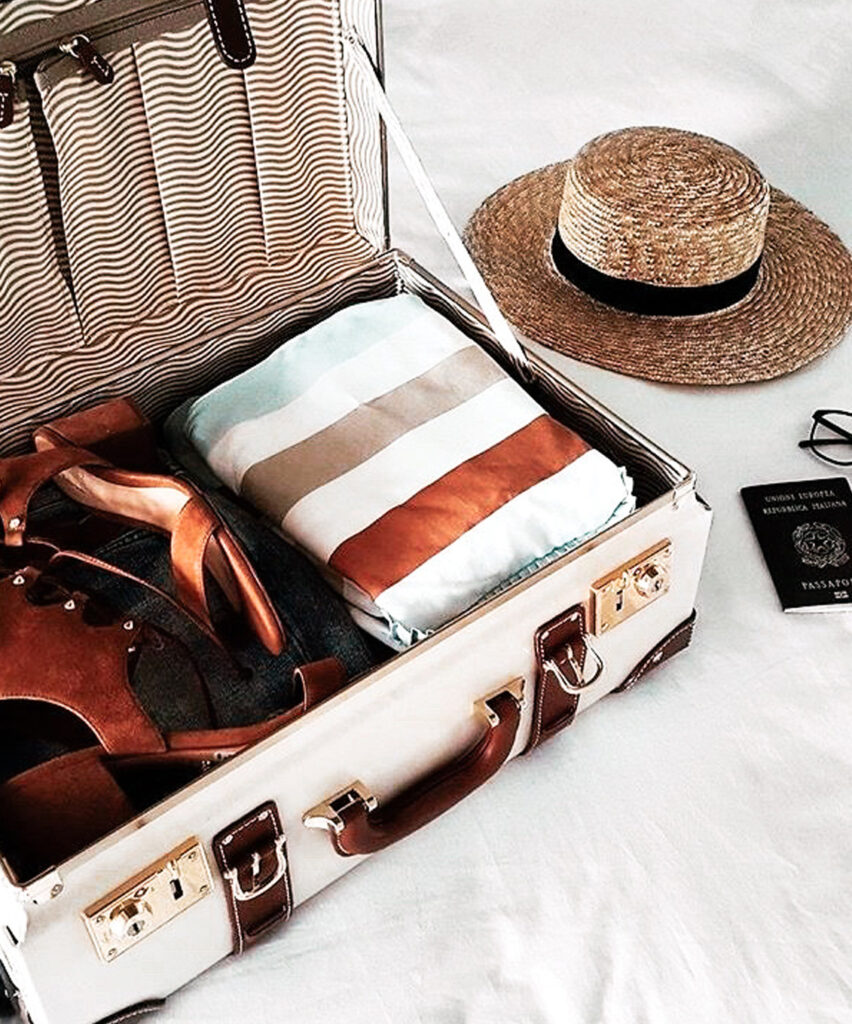 Valise ouverte contenant des vêtements, des sandales et des accessoires de voyage, accompagnée d’un chapeau de paille.