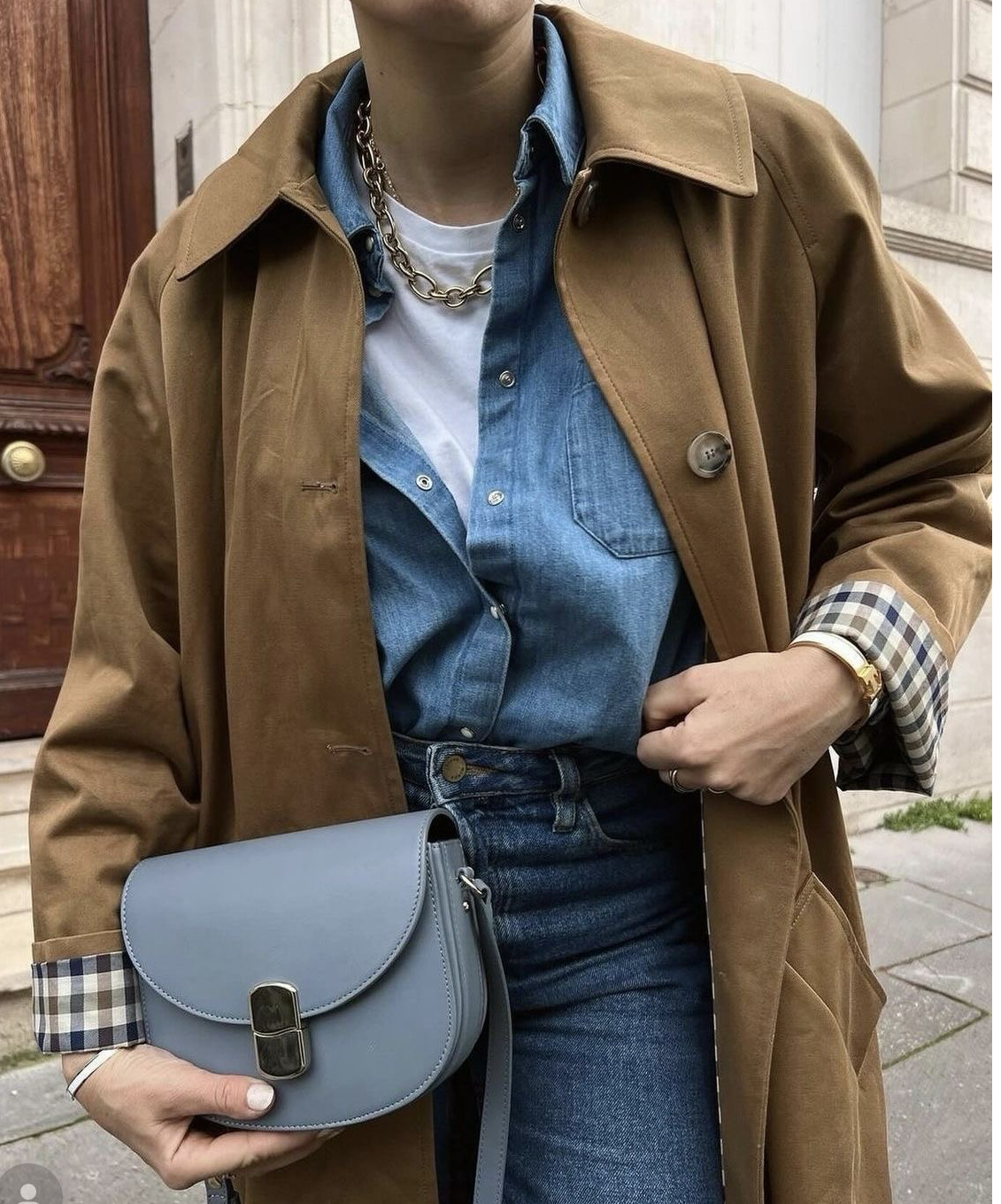 Femme portant un trench camel superposé à une chemise en denim, un t-shirt blanc et un jean, avec un sac bleu.
