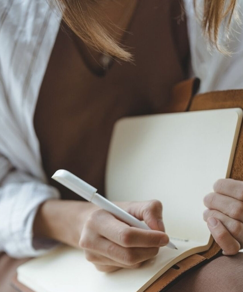 Femme écrivant dans un carnet avec un stylo, accompagnée d’une tablette.